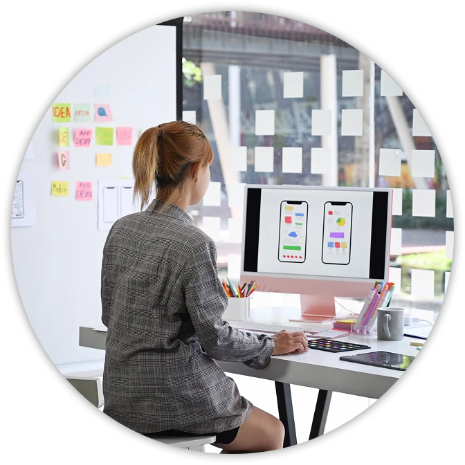 A person with light brown hair in a ponytail sits at a modern desk, looking at a large desktop monitor that displays two colorful mobile app wireframes. Sticky notes are arranged on the glass wall beside them, and design tools, a keyboard, and a tablet are spread across the clean white workspace in a bright office.