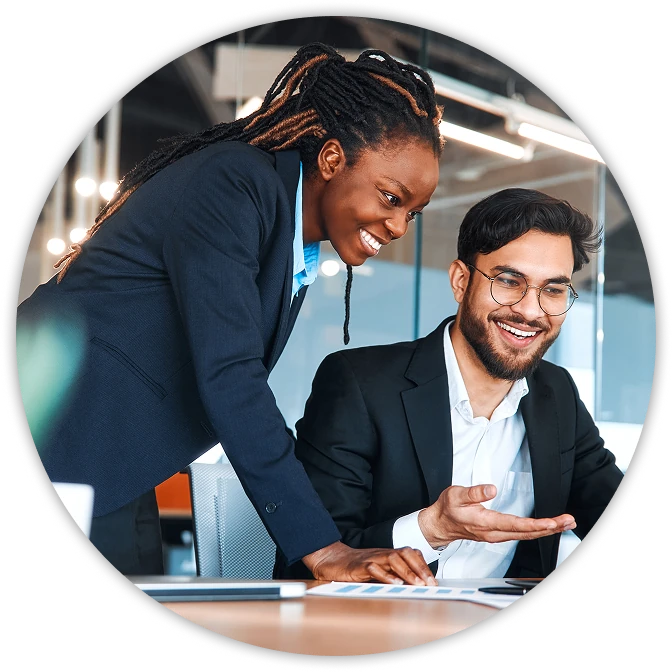 Two coworkers in an office collaborate at a desk; one leans over and points while the other, wearing glasses, smiles and gestures toward a laptop and printed charts.