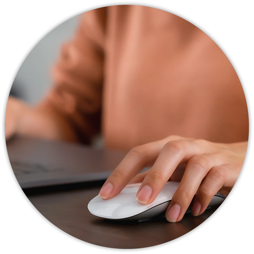 A person’s hand resting on a white computer mouse next to a laptop, suggesting someone working at a computer.