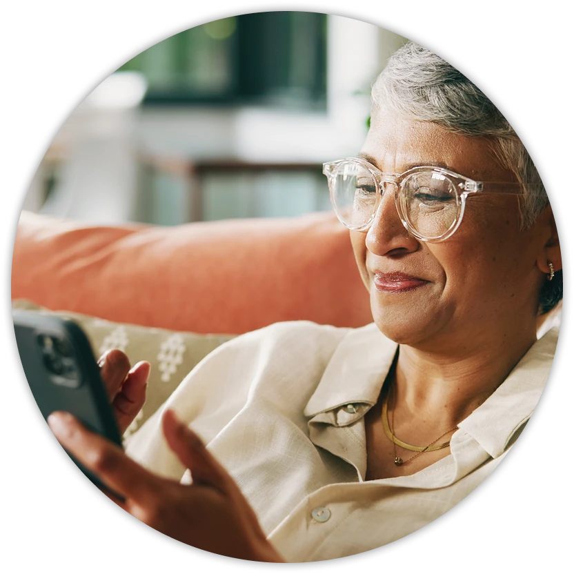 Older woman with short gray hair and clear glasses relaxing on a couch, smiling as she looks at her smartphone.