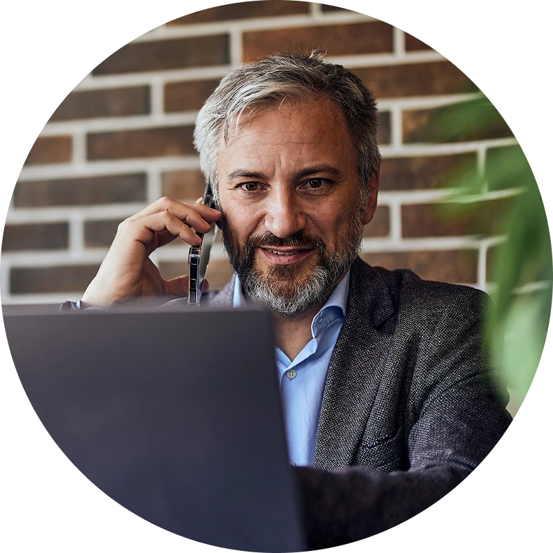 Middle-aged man with gray hair and beard sitting at a laptop, smiling while talking on a smartphone in front of a brick wall.