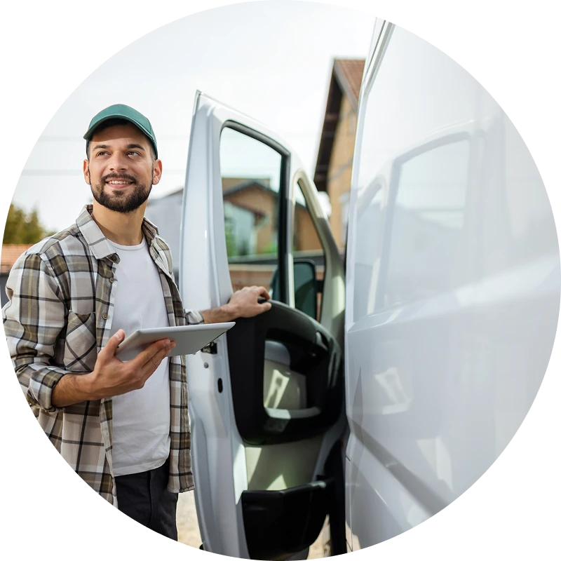 Smiling man wearing a green cap and plaid shirt holds a tablet while standing beside an open white van door, looking off to the left outdoors.